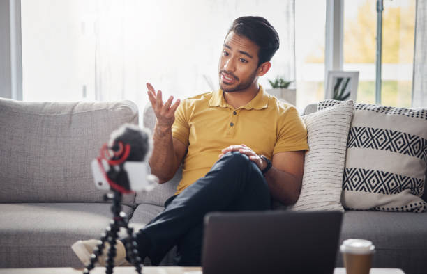 istock Content creator, laptop and phone, Indian man on sofa recording webinar, broadcast and live streaming in home. Internet, webcam and online influencer talking with equipment for social media channel. 1618257741