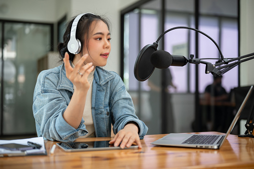 A professional Asian female radio host or podcaster is speaking into a microphone, announcing news and broadcasting her live audio podcast in the studio.