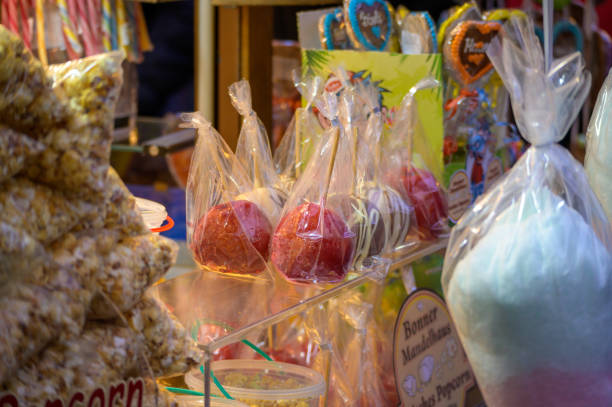 Bonn, Germany - December 5, 2022: Candied and chocolate covered apples wrapped in plastic amongst popcorn and cotton candy at a Christmas market stall