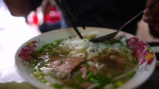 A man using chopsticks and spoon eating traditional Pho Bo vietnamese soup with beef and rice noodles on a metal table