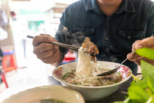 A man using chopsticks and spoon eating traditional Pho Bo vietnamese soup with beef and rice noodles on a metal table