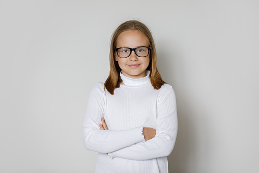 Cheerful smart confident successful child girl in glasses standing with crossed arms on white wall background, studio portrait