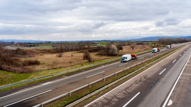 3台の白いミニバンが、古い高速道路と曇りの天候で農業機械の一部を輸送しています。 - car delivering white mini van ストックフォトと画像