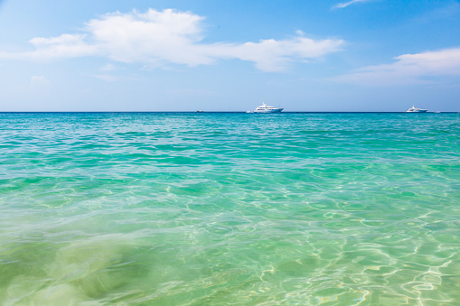 A tropical ocean scene with turquoise water and white yachts. The ocean water is a beautiful turquoise color and is crystal clear. The sky is a bright blue with a few wispy clouds. There are two white yachts in the distance on the horizon. The water is calm and the sun is shining brightly, creating a peaceful and serene mood.