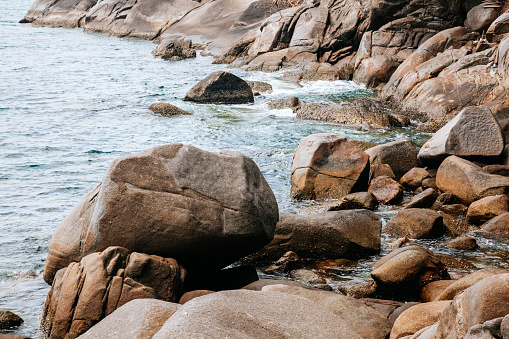 A rocky coastline with large boulders and rocks. The rocks are various shades of brown and gray. The water is a deep blue-green color and is crashing against the rocks. The sky is not visible in the image. The mood of the image is peaceful and serene.