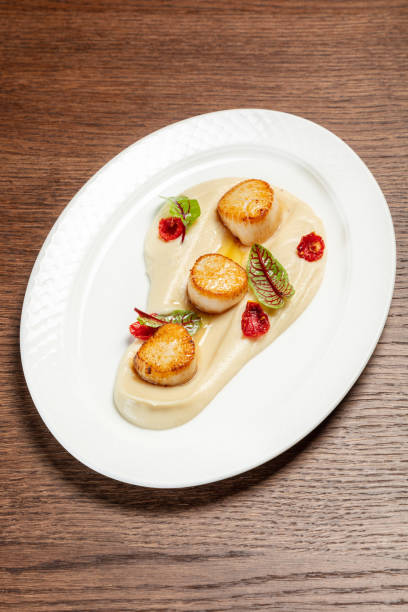 istock Fried sea scallop with mashed potatoes, lettuce and paprika slices on a light ceramic plate. The dishes are on a brown, wooden countertop. 1618494545