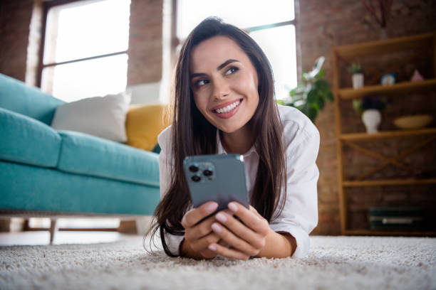 istock Photo of minded lady lying on carpet use cell smart gadget apple iphone pro max thinking order web new purchase for new apartment 1618499993