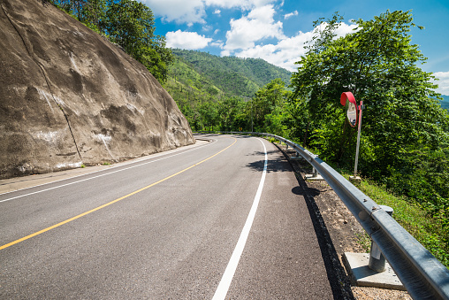 Downhill road turning with crash barrier, convex mirror and landslide concrete wall protection on the mountains in blue sky sunny day. Transport and traffic concept.