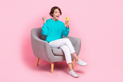 Full size photo of pretty young girl sit armchair raise fist hold telephone dressed stylish blue outfit isolated on pink color background.