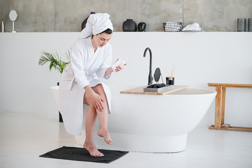 Woman in bathrobe and towel applying body lotion or balm on legs. Skincare after shaving, epilation, depilation or hair removal. Medical gel for treatment of varicose veins disease or edema.