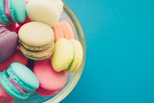 Colorful French macaroons in glass bowl on blue background copy space. Dessert food, design concept.