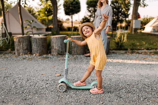 Little cute girl learning how to ride a push scooter while her mother helping her. Mother and daughter having fun with push scooter at camping.