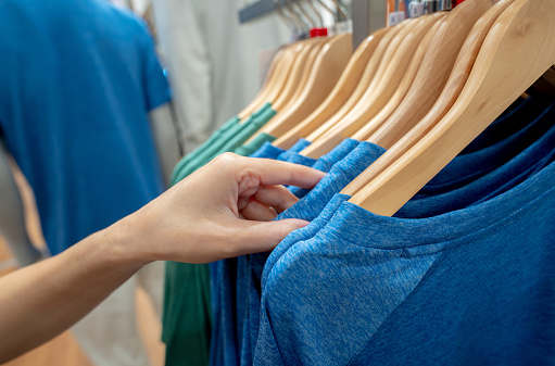 Woman shopping shirt in clothing store. Woman choosing clothes. Shirt on hanger hanging on rack in clothing store. Fashion retail shop inside shopping mall. Clothes on hangers in a clothes shop.
