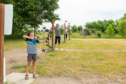 Young boy and his two older sisters practicing archery at an outdoor archery range on a cloudy summer day. They all have their bows drawn and are aiming at targets which are not visible in this image.