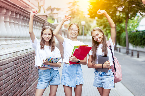 Three happy teenage girls (14-15 years old) walk to school, holding books and notebooks. Their faces light up with excitement for the first day of school and the beginning of a new academic year. Young females rejoice in passing exams.