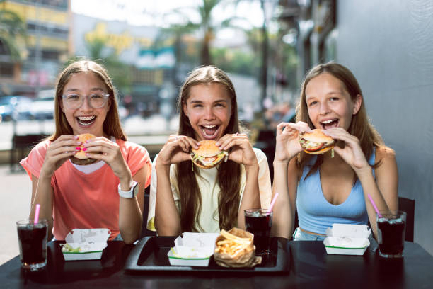 Fast food restaurant. Three joyful smiling teenage girls (14-15 years old) sitting at a cafe outdoors, enjoying hamburgers and cola (coke) fresh drinks. Young females holding burgers, ready to eat them with pleasure. Teenagers eating at fast food restaurant. spanish hamburger stock pictures, royalty-free photos & images