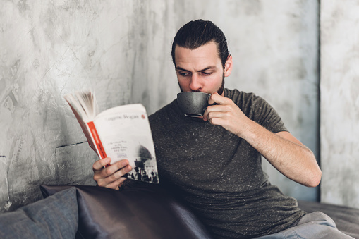 Handsome bearded hipster man relaxing holding and reading book with coffee while sitting on chair in cafe