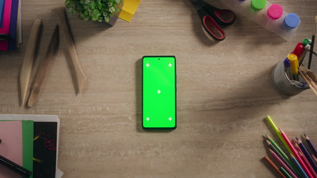 Top Down View of a Smartphone with Mock Up Green Screen Chromakey Display with Motion Tracker Placeholders. Static Footage of a Cellphone Device on a Table with Designer Art Supplies and Craft Tools