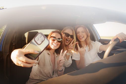 Happy young women in casual clothes with stylish eyeglasses sitting in car and smiling with v fingers while taking selfies on smartphone and looking at screen