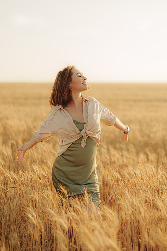 Positive young female in casual gown and shirt standing in dry grain field with stretched arms while enjoying vacation in countryside on sunny day