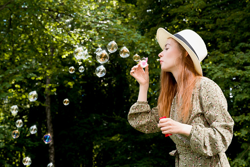 Side vie on a girl with red hair blowing bubbles in the park in summer wearing dress and a hat