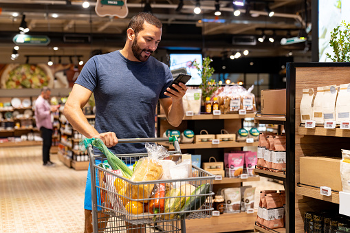 Fit Latin American man following a shopping list while buying healthy groceries at the supermarket