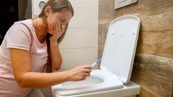 Young woman looking on positive pregnancy test sitting on toilet and holding her sickness. Intoxication and nausea during pregnancy.