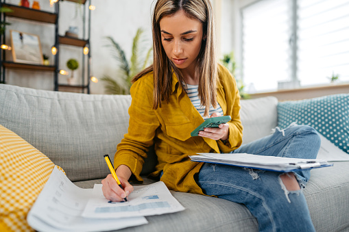 Beautiful young woman checking her finances using smartphone while sitting on the sofa.