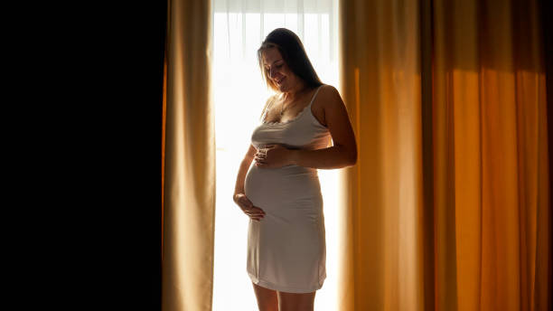 silhueta de linda mulher grávida sorridente parada na grande janela do quarto. conceito de gravidez feliz e esperando um bebê - camisola - fotografias e filmes do acervo