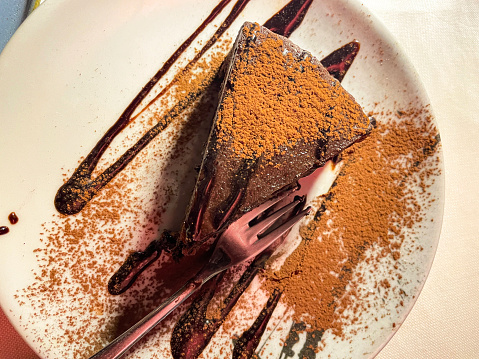 Stock photo showing close-up, elevated view of a white plate containing a dessert fork and a slice of chocolate tart with crispy, fluted pastry crust, dusted with cocoa powder and drizzled with a chocolate sauce.