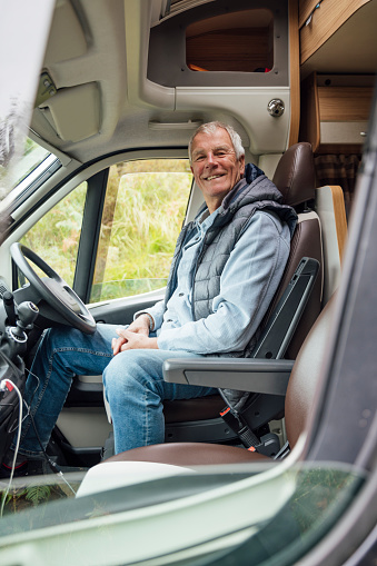 An senior man is sitting in the driving seat of his camper van. He is smiling at the camera. He is looking towards the passenger door and looks like he is ready off on his road trip.