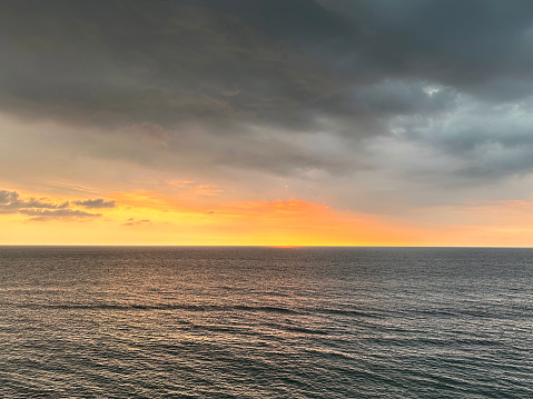 Stock photo showing a moody, dramatic orange sky as sun sets below grey bank of cloud on the horizon over the sea.