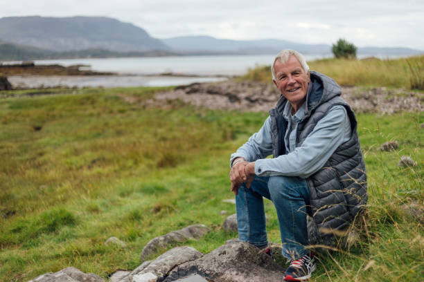 A man sits on a rocky perch in the Scottish Highlands, taking in the breathtaking views. Behind him is Loch Torridon. He is wearing a sleeveless, hooded puffer jacket.
