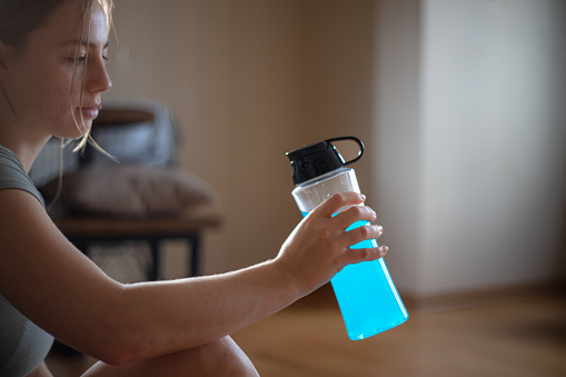 Young blonde woman is holding energy drink bottle in living room at home.