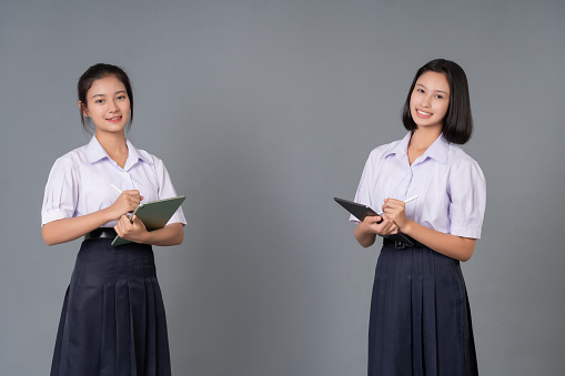 Two teenage Asian female students with short hair and long hair. wear school uniform Hand holding laptop and pen for homework, smiling brightly, photographed in gray background studio