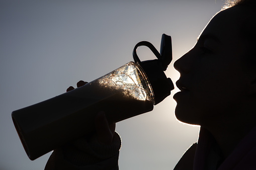 Young blonde lady is drinking smoothy after outdoor exercise at sunset.