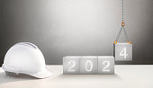 Happy New Year 2024 Construction and Industry. White Helmet and a numbered concrete cube on the desk of an engineer or construction worker. building with Cranes construction concept.
