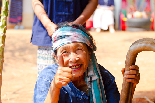 Happy looking and gesturing rural thai senior woman with headscarf and a wooden stick is sitting on floor and ground in rural village and town in north Thailand and Chiang Mai province. Woman is wearing traditional blue farmer fashion. Woman is gesturing and smiling spontaneously by herself into camera. No request for posing or staged gesture, pure natural reaction of friendly old woman happy being photographed. Behind woman another farm woman is standing partially in frame