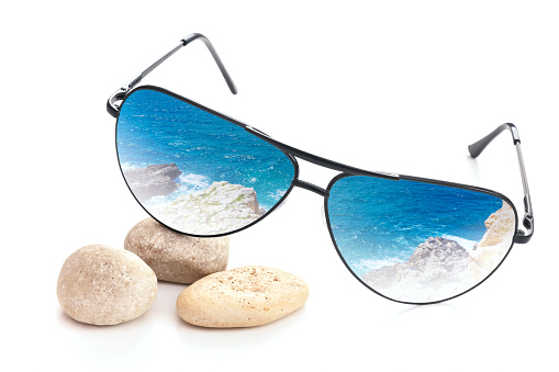 Sunglasses with blue summer sea reflection in lens. Glasses on sea pebbles stones on white background.