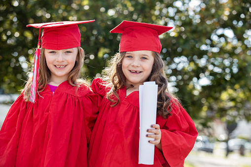 Beautiful little girls graduation from preschool and are ready for kindergarten. Students are wearing graduation caps and gowns, and holding diplomas, Children are standing outside of elementary school