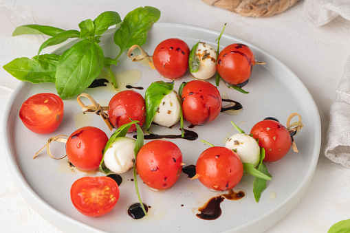 Caprese skewers with tomatoes, mozzarella balls, basil and spices on white table, closeup.  Italian homemade food and a healthy diet concept.