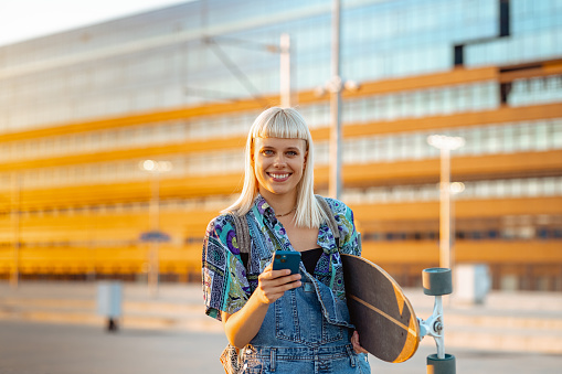 Young woman with blond hair holding a longboard on the street. She is looking at the camera