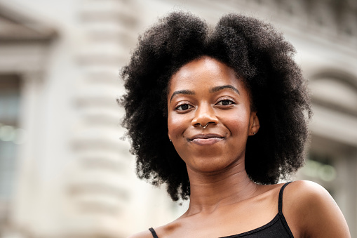 Head-shot of happy black curly woman outdoors. Blurry background, she is looking at camera. Positively concept, happiness and good vibes concept.