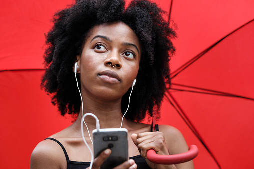 Black curly woman under red umbrella is listening to music on headphones connected to a mobile phone. She has a nose piercing and she is looking away focused in music.