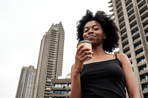 Portrait of smiling black curly woman drinking a cup of coffee surrounded by high skyscrapers