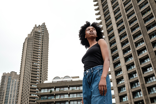 Serious black curly woman surrounded by skyscrapers is looking away in a proud and confident way. Barbican London vibes.