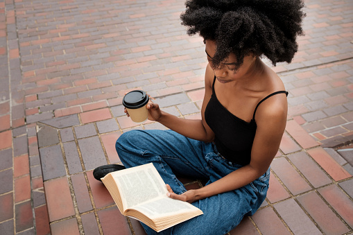 Curly black woman is reading a book and drinking a cup of coffee. She is sitting on the tiled red floor outdoors.