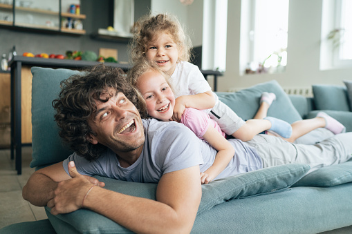 Photo of a young father with two kids lying on the top of each other being cheerful and playful. Single father with his two little children playing on the sofa at home.
