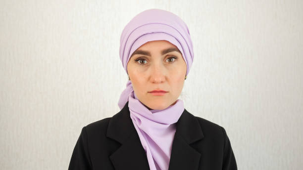 Muslim woman in pinkish hijab looks with depressed and upset expression. Sad lady wearing black blazer at white studio wall