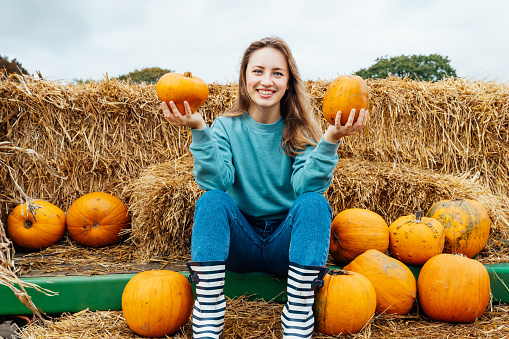 Smiling young woman sitting on straw bales and holding pumpkins. selecting best one for Thanksgiving and Halloween holidays decoration on agriculture farm. Pumpkin harvest. Autumn fall festive mood.